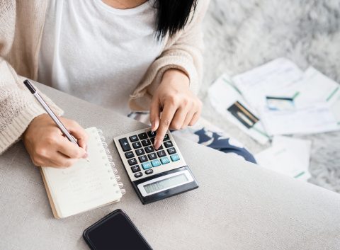 Person using a calculator and writing in a notebook, with bills and documents scattered on the floor in the background.
