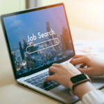 Person typing on a laptop with a “Job Search” screen displayed, showing a cityscape background.