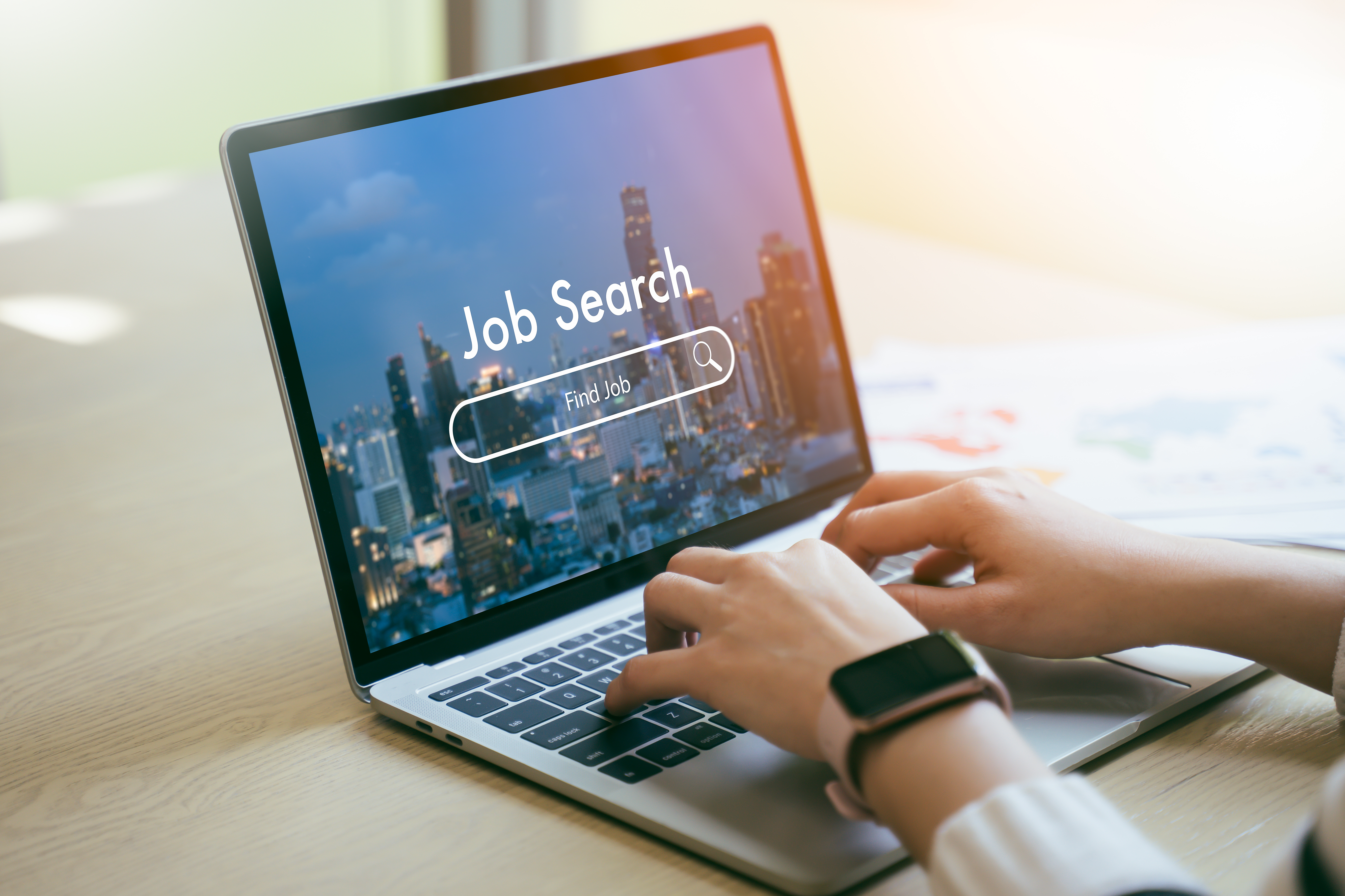 Person typing on a laptop with a “Job Search” screen displayed, showing a cityscape background.