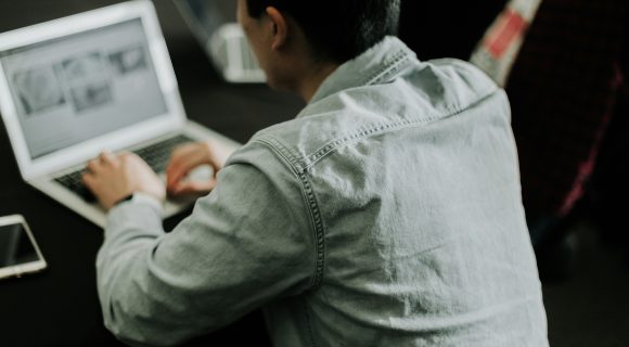 Person in a denim shirt working on a laptop at a dark table, viewed from behind.