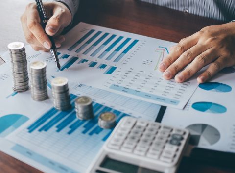 Person analyzing financial data with charts and stacked coins on a desk.