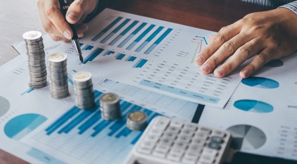 Persona analizando datos financieros con gráficos y monedas apiladas sobre un escritorio.