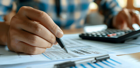Close-up of a person analyzing financial documents with a pen in hand and a calculator nearby on a desk.