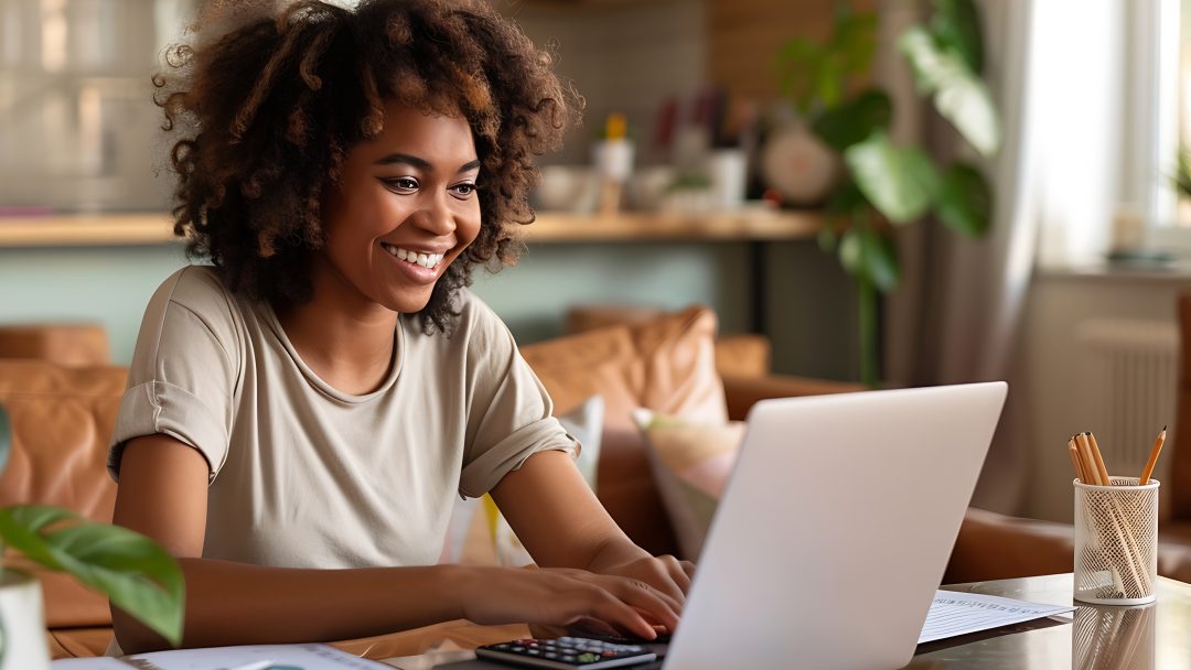 A smiling woman working on a laptop at home with charts and a calculator on the table.