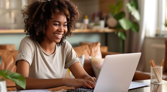 A smiling woman working on a laptop at home with charts and a calculator on the table.