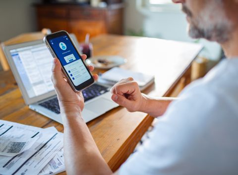Person reviewing digital banking app on mobile phone with bills, receipts, and an open laptop on a wooden desk.