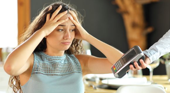 A worried person throws their hands on her head as a restaurant server extends a payment terminal toward them to pay the bill.