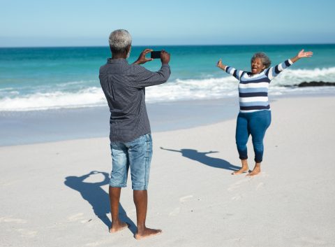Vista frontal de una pareja afroamericana de pie en la playa con el cielo azul y el mar de fondo, la mujer levanta las manos y sonríe, mientras el hombre le hace una foto con su smartphone.