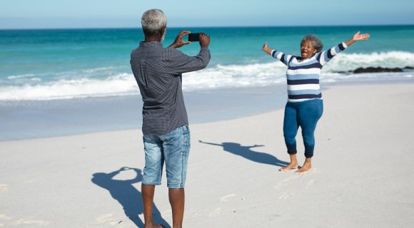 Front view of an African American couple standing on the beach with blue sky and sea in the background, the woman raising her hands and smiling, while the man takes a photo of her with his smartphone