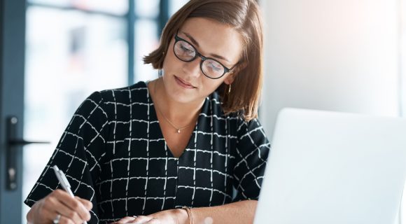 Mujer profesional escribiendo en un cuaderno mientras trabaja con un portátil en una oficina moderna.
