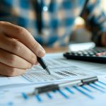 Close-up of a person reviewing financial documents with a pen and calculator on a desk.