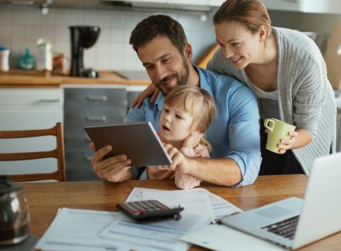 A happy family sitting at the kitchen table, reviewing financial documents on a tablet, with a laptop and calculator nearby.