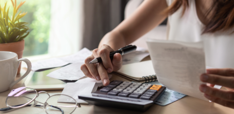 A woman calculating expenses at a desk, holding a receipt while using a calculator, with financial documents and a notebook in front of her.