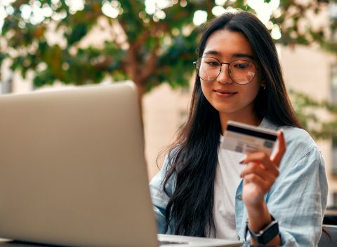 A young woman wearing glasses and a smartwatch holds a credit card while using her laptop outdoors.