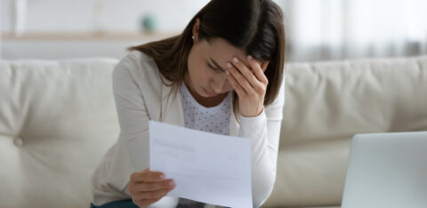 A worried woman sitting on a couch, holding a bill in one hand and resting her head on the other, surrounded by financial documents and a laptop.