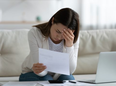 A worried woman sitting on a couch, holding a bill in one hand and resting her head on the other, surrounded by financial documents and a laptop.