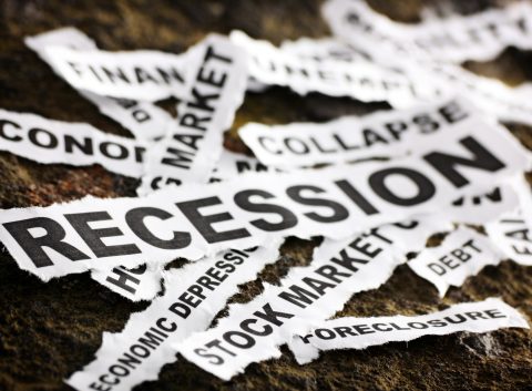 Recortes de periódico rotos con palabras relacionadas con la crisis económica como "Recesión", "Bolsa", "Colapso" y "Deuda" esparcidas sobre una superficie rugosa.