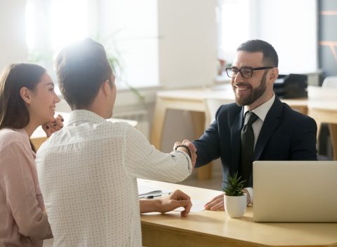 Un sonriente asesor financiero vestido de traje estrecha la mano de un cliente masculino, mientras una clienta le observa sonriente en una moderna oficina.