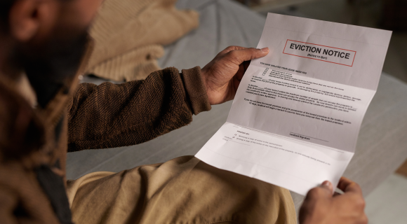 A person wearing a brown sweater and beige pants sits on a bed, holding and reading an eviction notice with a serious expression.