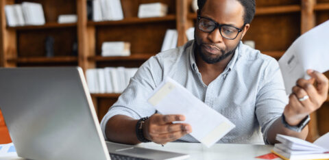 A man wearing glasses sits at a desk with a laptop, reviewing financial documents and bills with a focused expression.