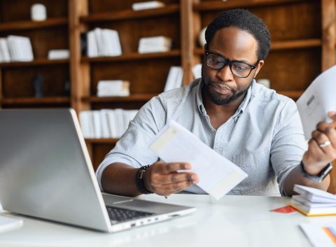 A man wearing glasses sits at a desk with a laptop, reviewing financial documents and bills with a focused expression.