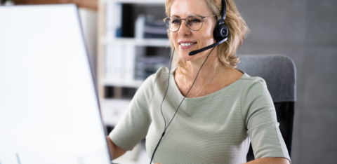 A smiling middle-aged woman wearing a headset works on her computer in a modern office setting.