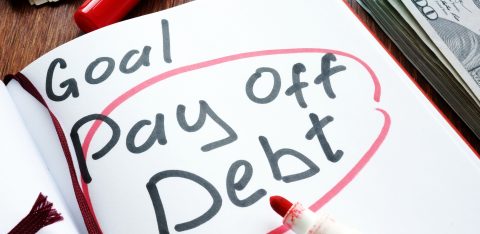 Notebook with the handwritten phrase “Goal: Pay Off Debt” circled in red marker, with cash and a red pen nearby.