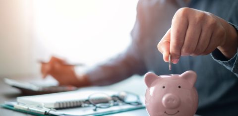 Person placing a coin into a pink piggy bank on a desk, with documents, a pen, and a calculator in the background.