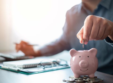 Person placing a coin into a pink piggy bank on a desk, with documents, a pen, and a calculator in the background.