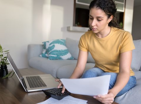 Young woman sitting on a couch reviewing financial documents with a calculator and laptop on the table.