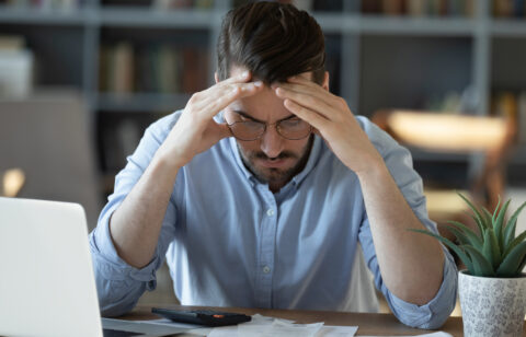 Stressed man sitting at a desk with his hands on his head, looking at financial documents and a calculator.
