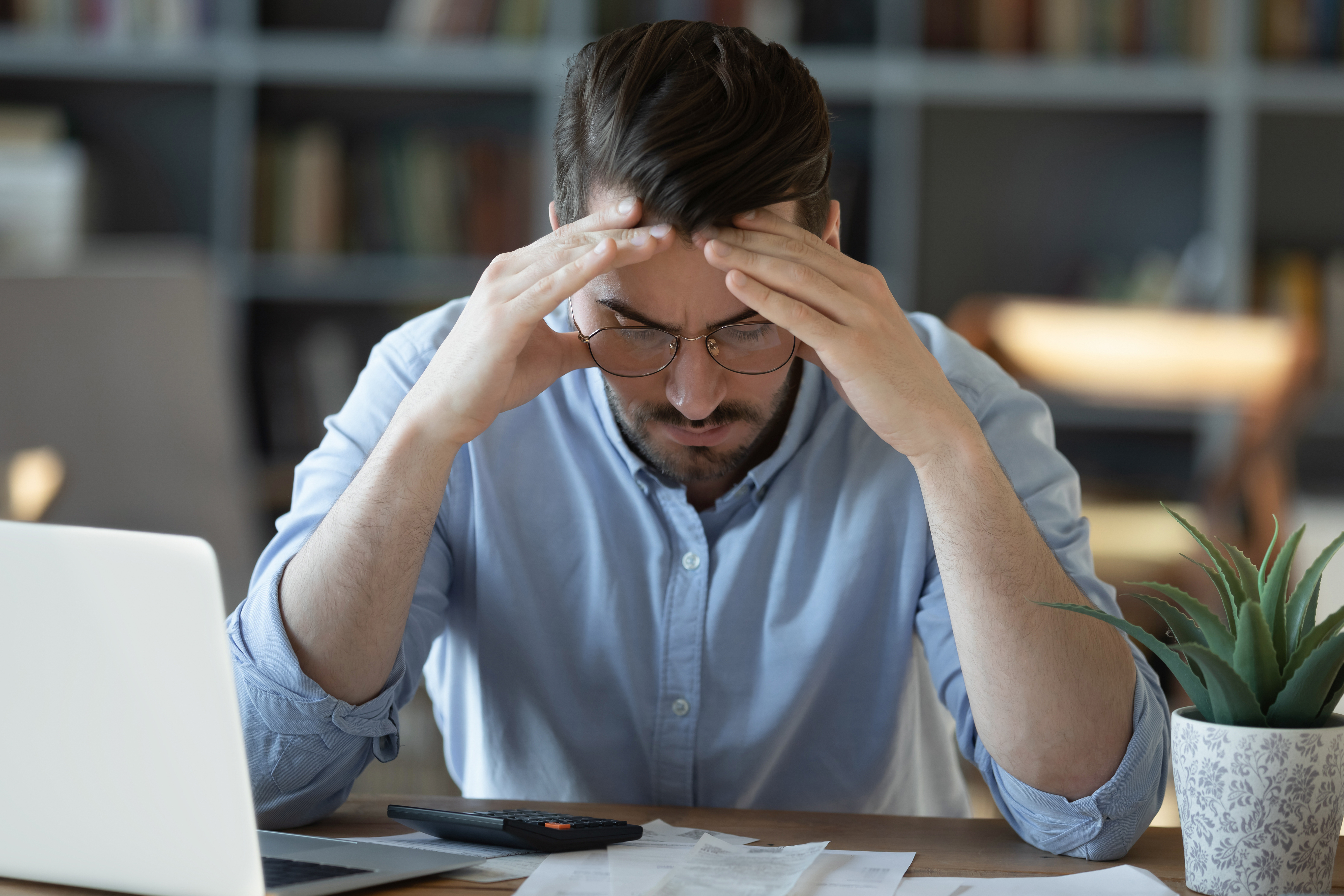 Stressed man sitting at a desk with his hands on his head, looking at financial documents and a calculator.