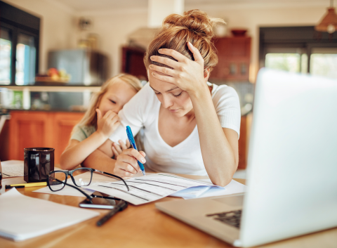 A stressed mother sits at a kitchen table, reviewing financial documents with a pen in hand, while her young daughter leans on her for comfort.