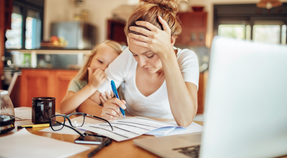 A stressed mother sits at a kitchen table, reviewing financial documents with a pen in hand, while her young daughter leans on her for comfort.