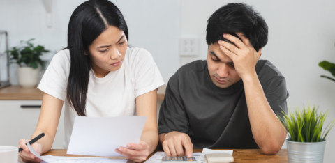 Stressed couple sitting at a table reviewing financial documents and using a calculator to manage expenses.