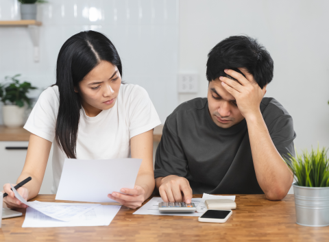 Stressed couple sitting at a table reviewing financial documents and using a calculator to manage expenses.