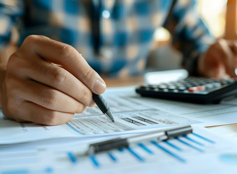 Hand holding a pen over printed financial documents with graphs, analyzing expenses and calculating figures.