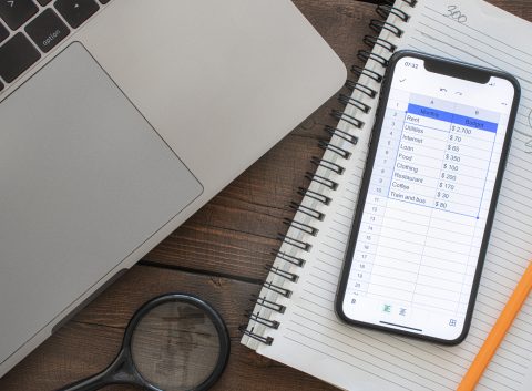 A smartphone displaying a digital budgeting spreadsheet next to a laptop, notebook, and pencil on a wooden desk.