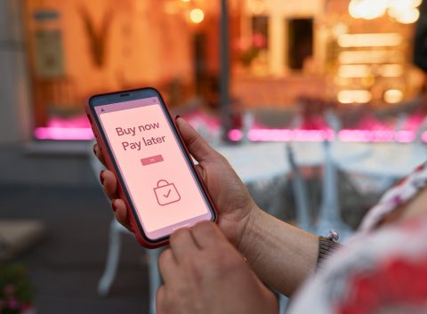 Person holding a smartphone displaying a "Buy now, pay later" message on a shopping app screen, with a blurred outdoor café background.