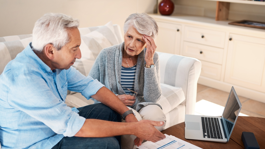 Older couple sitting on a couch reviewing financial documents with a laptop, looking concerned while discussing their finances.