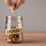 Hand placing a coin into a glass jar labeled “Emergency,” symbolizing saving money for unexpected expenses.
