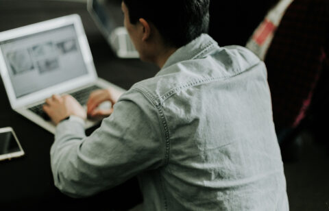 Over-the-shoulder view of a man typing on a laptop with a smartphone on the table beside him.