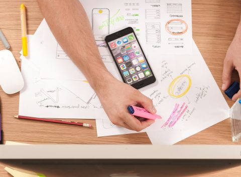 Person highlighting notes on wireframe sketches at a cluttered desk with a smartphone and colorful pens.