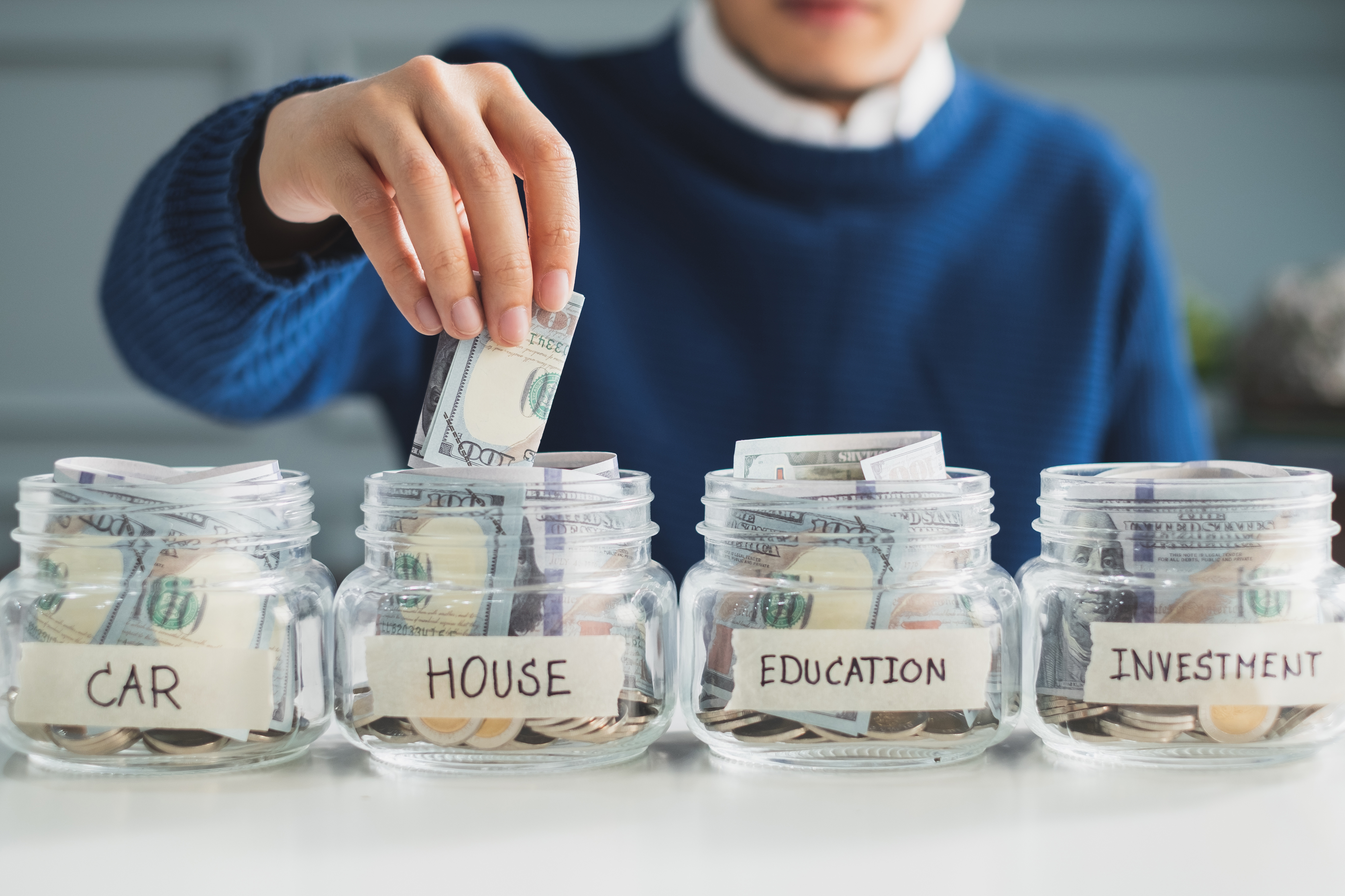 Person placing cash into a labeled savings jar for a house, alongside jars for a car, education, and investment.