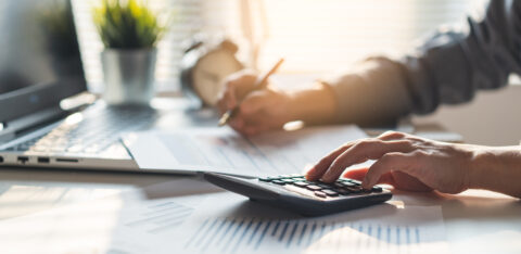 Person using a calculator and reviewing financial documents at a desk, with a laptop and charts in a sunlit office.