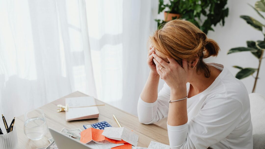 Stressed woman sitting at a desk with her head in her hands, surrounded by receipts, bills, and a calculator.