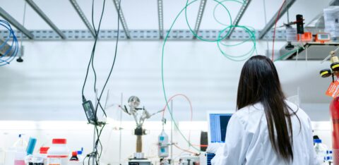 Researcher in a white lab coat analyzing data on a computer in a modern lab filled with wires, tubes, and lab tools.