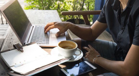 Person working on a laptop at an outdoor café table with a cup of coffee, a smartphone, and a notebook nearby.