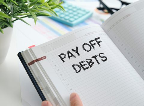 Close-up of an open planner with the handwritten words “Pay Off Debts” on a dated page, surrounded by office supplies.