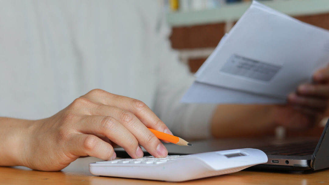 Close-up of hands using a white calculator and holding an envelope, with a pencil and laptop on a wooden desk.